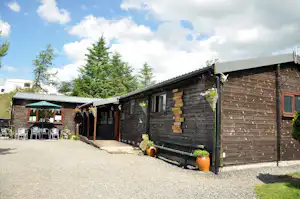 Holzblockhütte mit Terrasse am Campingplatz, Kiesweg, Töpfe mit Blumen und Bäume vor dem Gebäude.