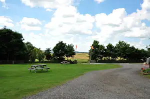 Grüner Campingplatz mit Rasenfläche, Kiesweg und Sitzbank, umgeben von Bäumen und Blick auf die Landschaft.