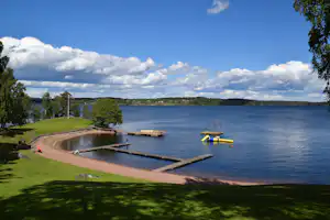 Seeufer mit Sandstrand, Steg und Schwimmplattformen, gelbem Schlauchboot im Wasser und Bäumen am Rand.
