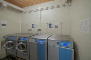 Waschraum mit mehreren Waschmaschinen und Wäscheautomaten an der Wand, Holzdecke und weiße Leitungen im Hintergrund.