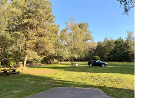 Grüner Campingplatz mit Rasenfläche, Baumreihe und einem geparkten Auto am Rand der Wiese.