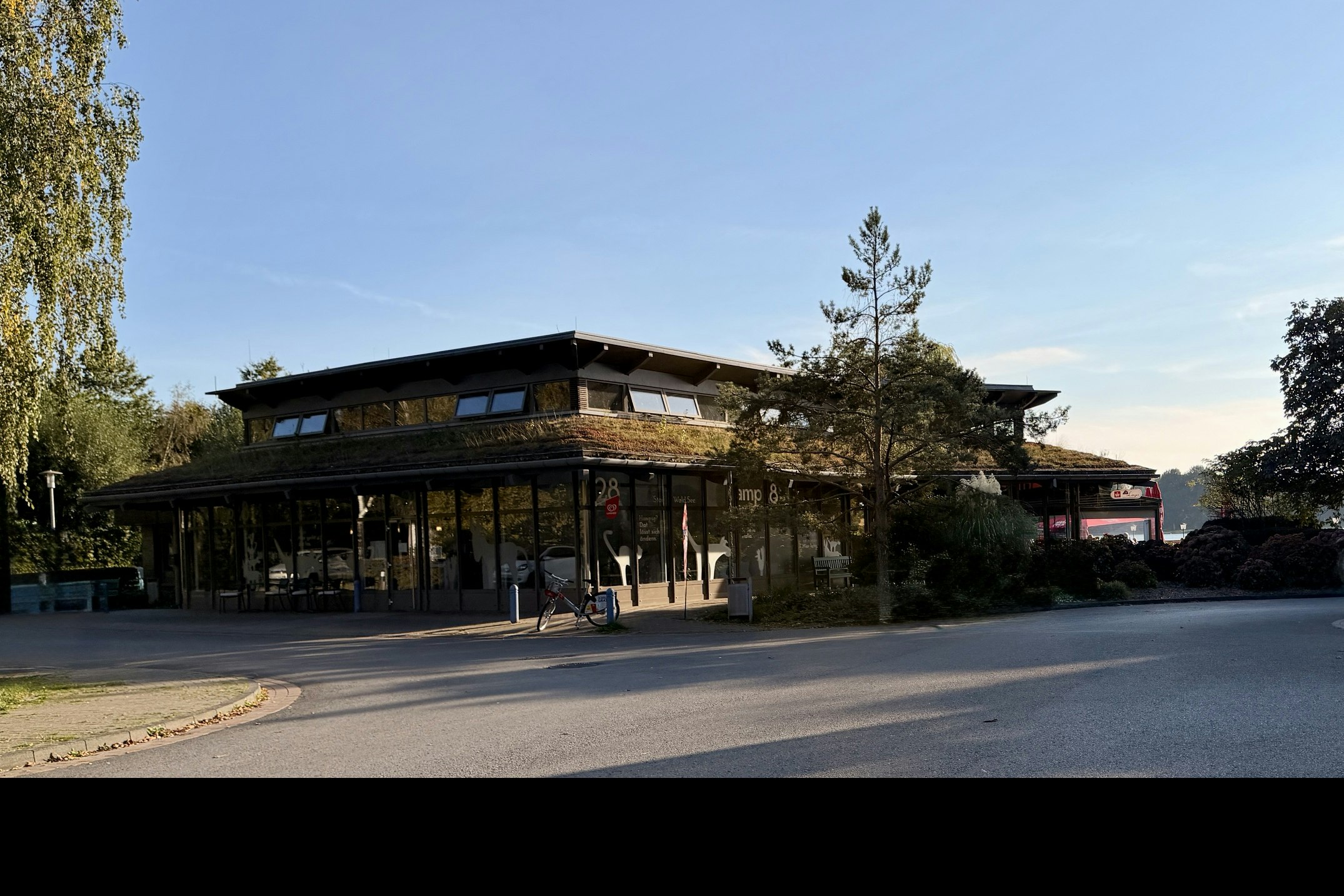 Campingplatz-Gebäude mit großer Glasfront, davor Straße und Bäume, im Hintergrund weitere Gebäude und ein Parkplatz.