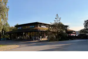Campingplatz-Gebäude mit großer Glasfront, davor Straße und Bäume, im Hintergrund weitere Gebäude und ein Parkplatz.