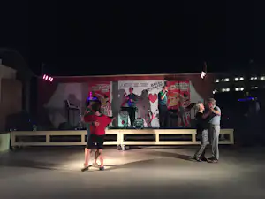 Abendliche Bühne auf dem Campingplatz mit Musikern, großen Bannern und zwei Personen vor dem Podium.