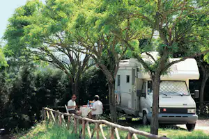 Wohnmobil auf dem Campingplatz neben einem Tisch unter Bäumen, zwei Personen sitzen am Rand des Stellplatzes.