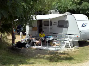 Wohnwagen mit Markise und Terrasse, Campingstühle und Tisch vor dem Eingang, umgeben von Bäumen und Grün.