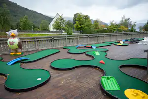 Minigolfanlage auf einer Holzterrasse mit grünen Bahnen, gelben Hindernissen und Blick auf Bäume und ein Gebäude im Hintergrund.