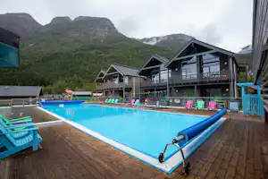 Schwimmbad im Campingbereich mit Holzterrasse, bunten Liegestühlen und Ferienhäusern vor Bergen und Wald im Hintergrund.
