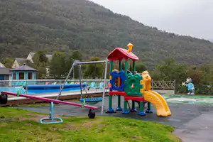 Bunter Spielplatz am Campingplatz mit Rutsche und Kletterelementen, daneben ein Pool und Rasen vor den Hügeln.
