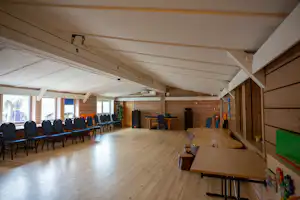 Überdachte Gemeinschaftshalle im Campingbereich mit langen Tischen, Stühlen, Holzdecke und großen Fenstern.
