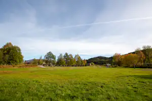 Weite grüne Campingwiese mit Bäumen, Feldweg und Blick in die Landschaft unter wolkigem Himmel.