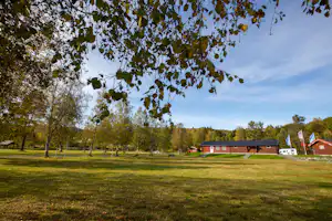 Weite Campingwiese mit Bäumen im Vordergrund und roten Gebäuden im Hintergrund, Blick in den Gartenbereich.
