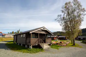 Holzchalet auf einem Campingplatz mit Veranda, Holzstufen und Weg, daneben weitere kleine Unterkünfte und Bäume.