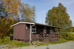Holzhütte mit Veranda und Treppe, seitlich ein Fenster, umgeben von Gras und Bäumen im Campingbereich.