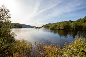 Ruhiger Fluss mit Uferbäumen und bewachsenem Rand, im Hintergrund Häuser und Hügel unter klarem Himmel.