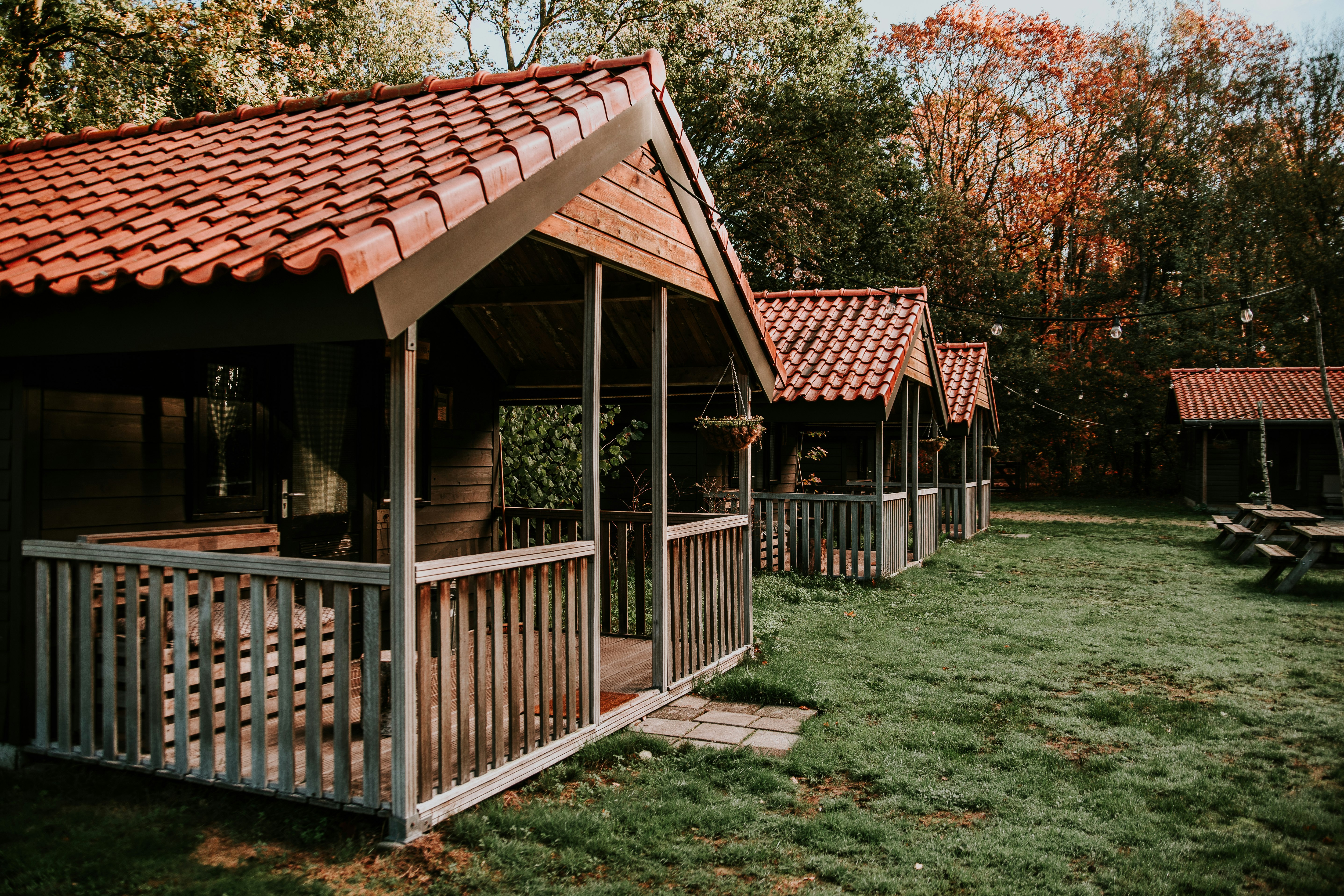Holzhütte mit überdachter Terrasse und Geländer, Rasenfläche daneben, im Hintergrund Bäume mit bunten Blättern.