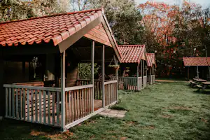 Holzhütte mit überdachter Terrasse und Geländer, Rasenfläche daneben, im Hintergrund Bäume mit bunten Blättern.