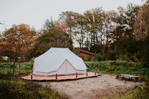 Weißes Zelt auf einem Holzpodest auf einem Campingplatz, umgeben von Bäumen und einem Kiesweg.