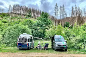 Stellplatz mit zwei Wohnmobilen, Fahrrad am Heck, Tisch und Stühlen im Grünen am Waldrand.