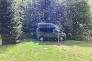 Camping-Van steht auf einer Wiese am Waldrand, umgeben von Bäumen und Hecken.