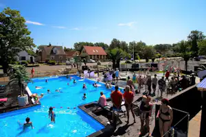 Großes Camping-Schwimmbad mit vielen Urlaubern im Wasser und auf Liegen, im Hintergrund Häuser und Bäume.