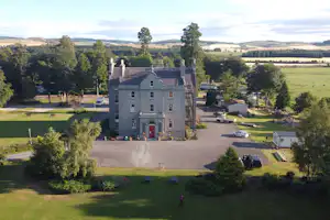 Landhaus mit gepflastertem Hof, Parkplatz und Bäumen, umgeben von Wiesen und Hügeln in ländlicher Camping-Umgebung.