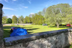 Campingzelt auf einer grünen Wiese, dahinter Bäume und weitere Zelte, im Vordergrund eine niedrige Steinmauer.