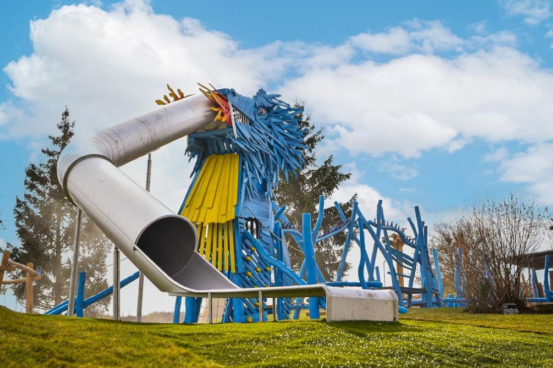 Blauer Spielplatz mit Rutsche und Klettergerüst, daneben ein weißer Rutschenturm und ein kleiner Sitzbereich auf Rasen.