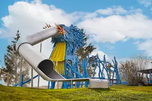 Blauer Spielplatz mit Rutsche und Klettergerüst, daneben ein weißer Rutschenturm und ein kleiner Sitzbereich auf Rasen.