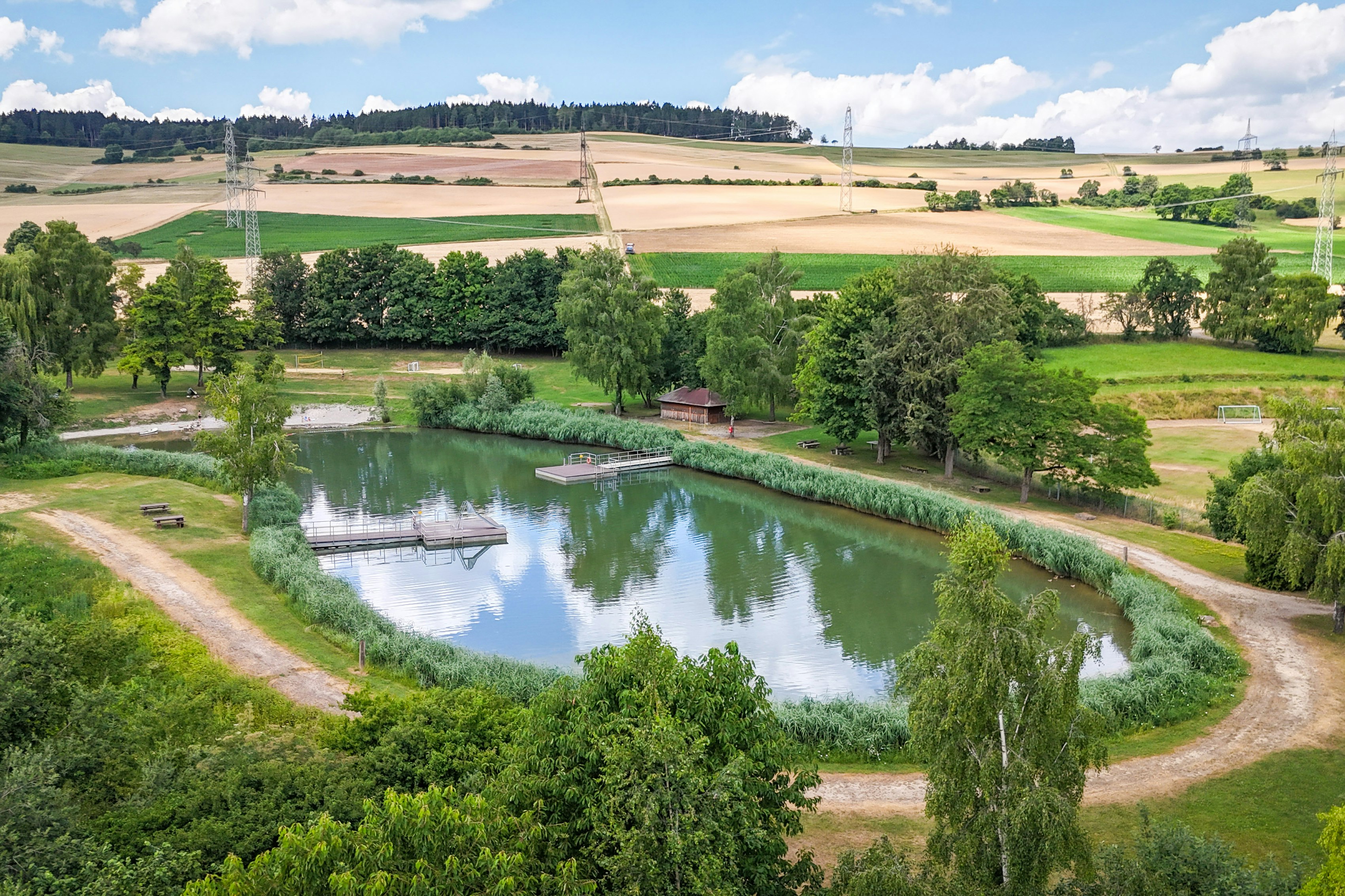 Ruhiger See mit Uferweg und Stegen, umgeben von grünen Bäumen und Feldern, Blick über die Landschaft.