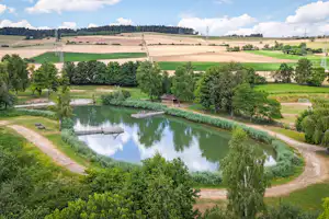 Ruhiger See mit Uferweg und Stegen, umgeben von grünen Bäumen und Feldern, Blick über die Landschaft.