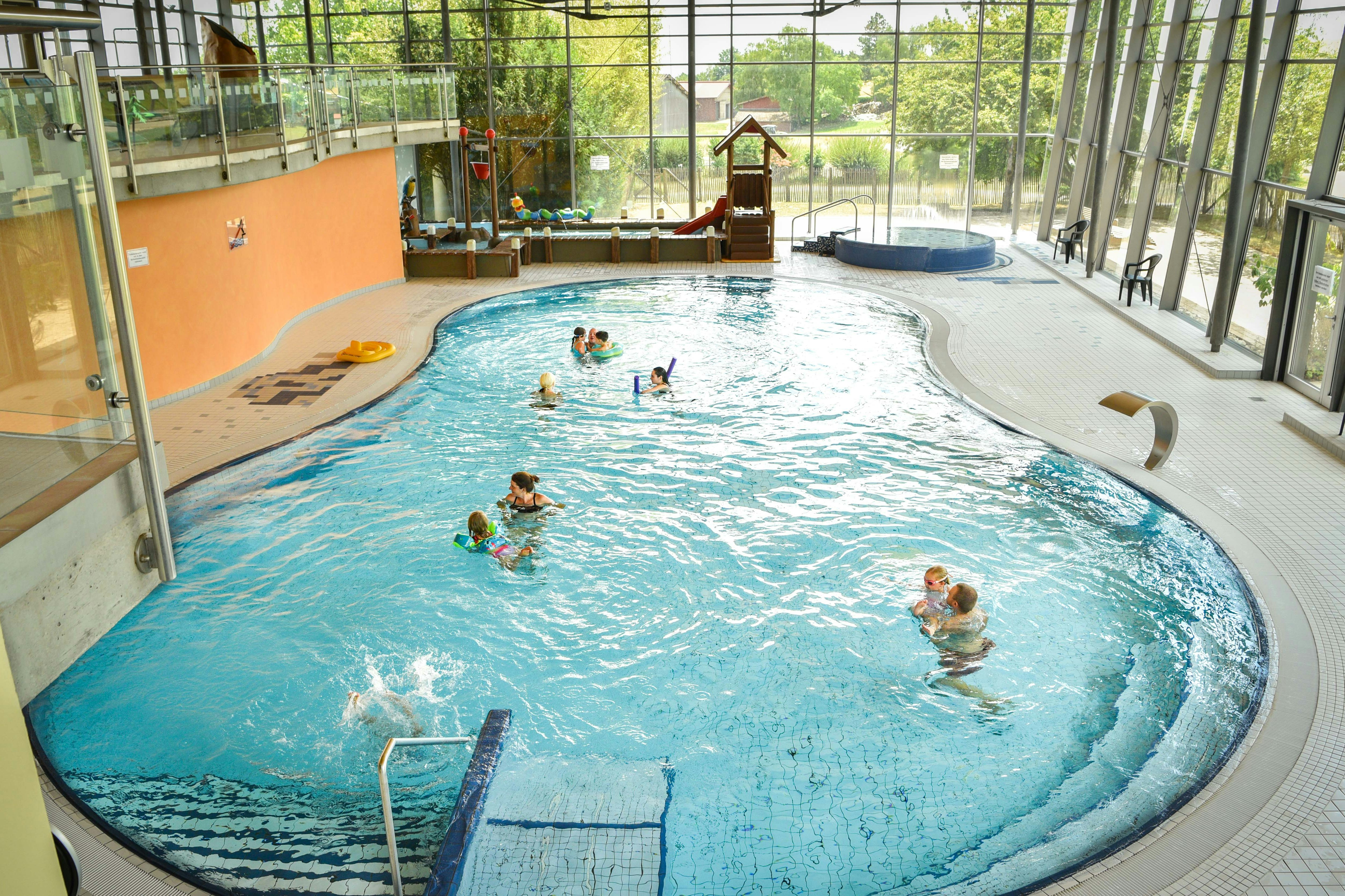 Innenpool mit Whirlpool-Ecke, mehrere Personen im Wasser und Liegen am Beckenrand, große Glasfront und Metallgeländer.