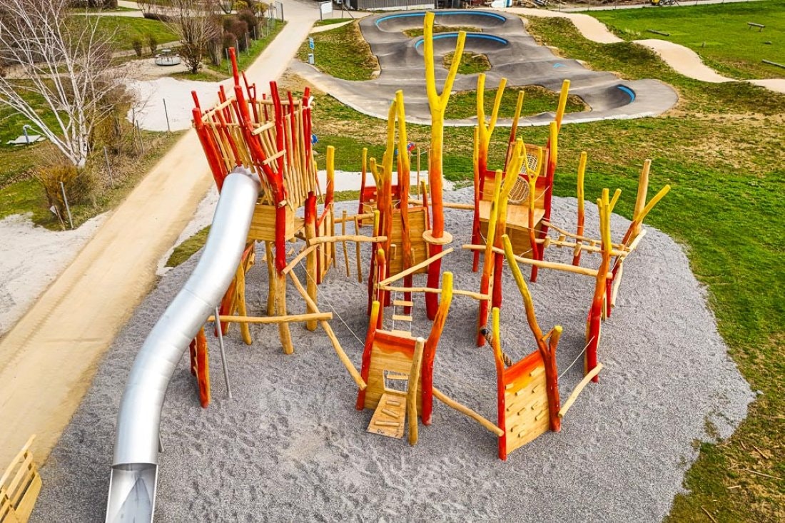 Spielbereich mit Klettergerüst aus Holz und gelben Stangen auf einem Kiesplatz, daneben Wege und Rasenflächen im Campinggelände.