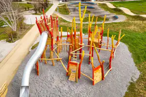Spielbereich mit Klettergerüst aus Holz und gelben Stangen auf einem Kiesplatz, daneben Wege und Rasenflächen im Campinggelände.