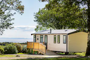 Ferienhaus mit Holzterrasse, großen Fenstern und Markise, umgeben von Bäumen; im Hintergrund ist der See zu sehen.