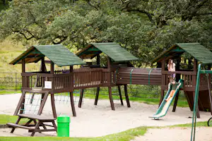 Spielplatz mit Sandfläche, Rutsche und Holz-Spielhaus mit grünen Dächern im Grünen, umgeben von Bäumen.