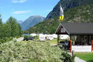 Campingplatz mit Wohnwagen, Flaggen am Eingang und Blick auf Berge und See, umgeben von Bäumen und Sträuchern.