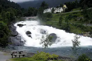 Wasserfall im Tal mit Blick auf Häuser am Hang, Steg und Bank am Ufer.