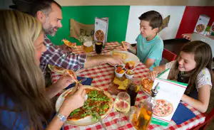 Familie am Camping-Tisch mit Pizza und Getränken, bunte Wand im Hintergrund, mehrere Teller und Besteck auf der Decke.