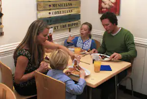Familie sitzt an einem Tisch im Campingbereich, isst und trinkt, im Hintergrund ein Schild mit Aufschrift „Musicians“.