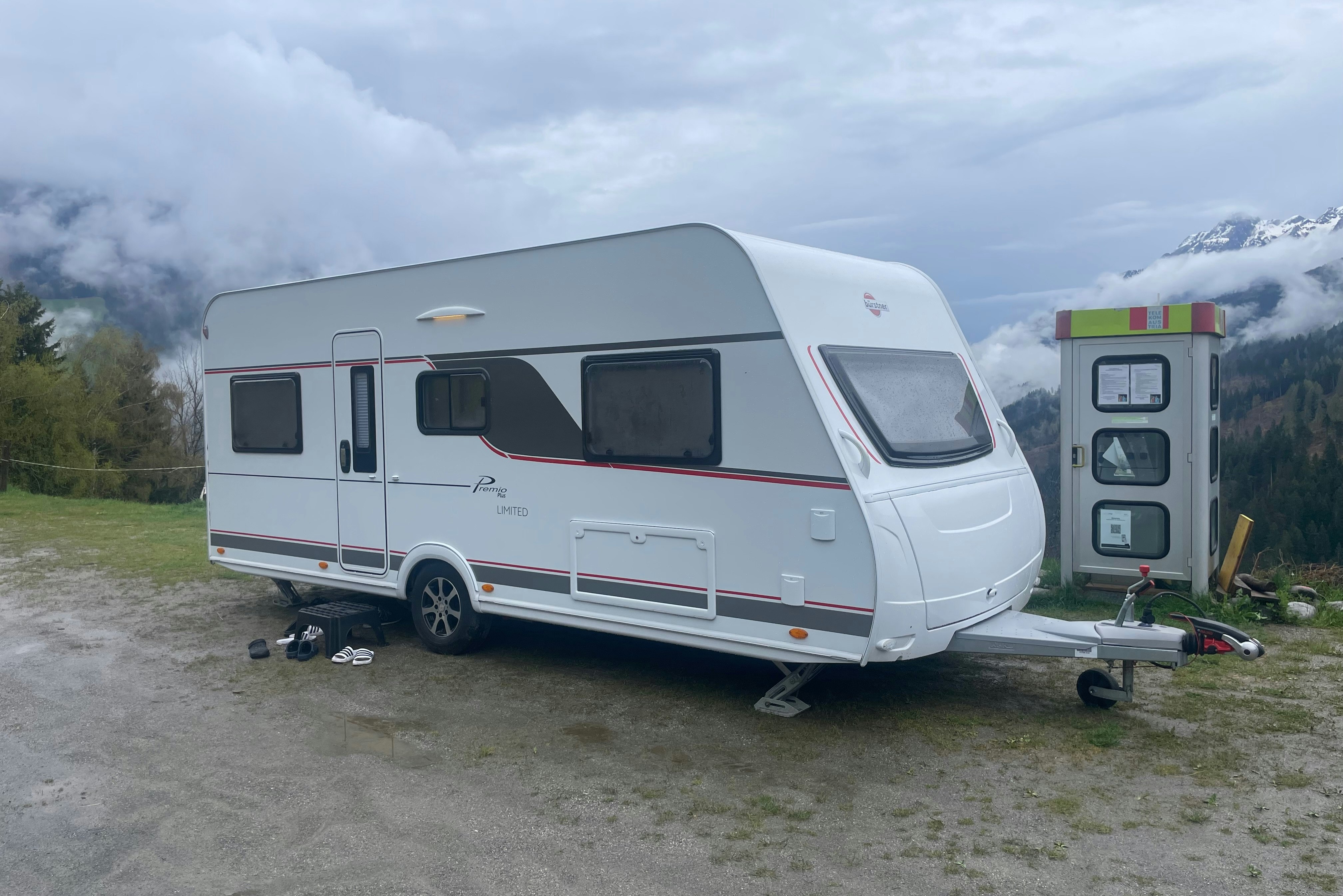 Weiße Wohnwagen auf einem Stellplatz, mit Vorzelt und Gepäck am Boden, im Hintergrund Berge und Bäume.