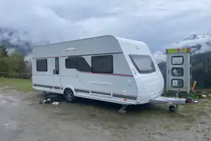 Weiße Wohnwagen auf einem Stellplatz, mit Vorzelt und Gepäck am Boden, im Hintergrund Berge und Bäume.