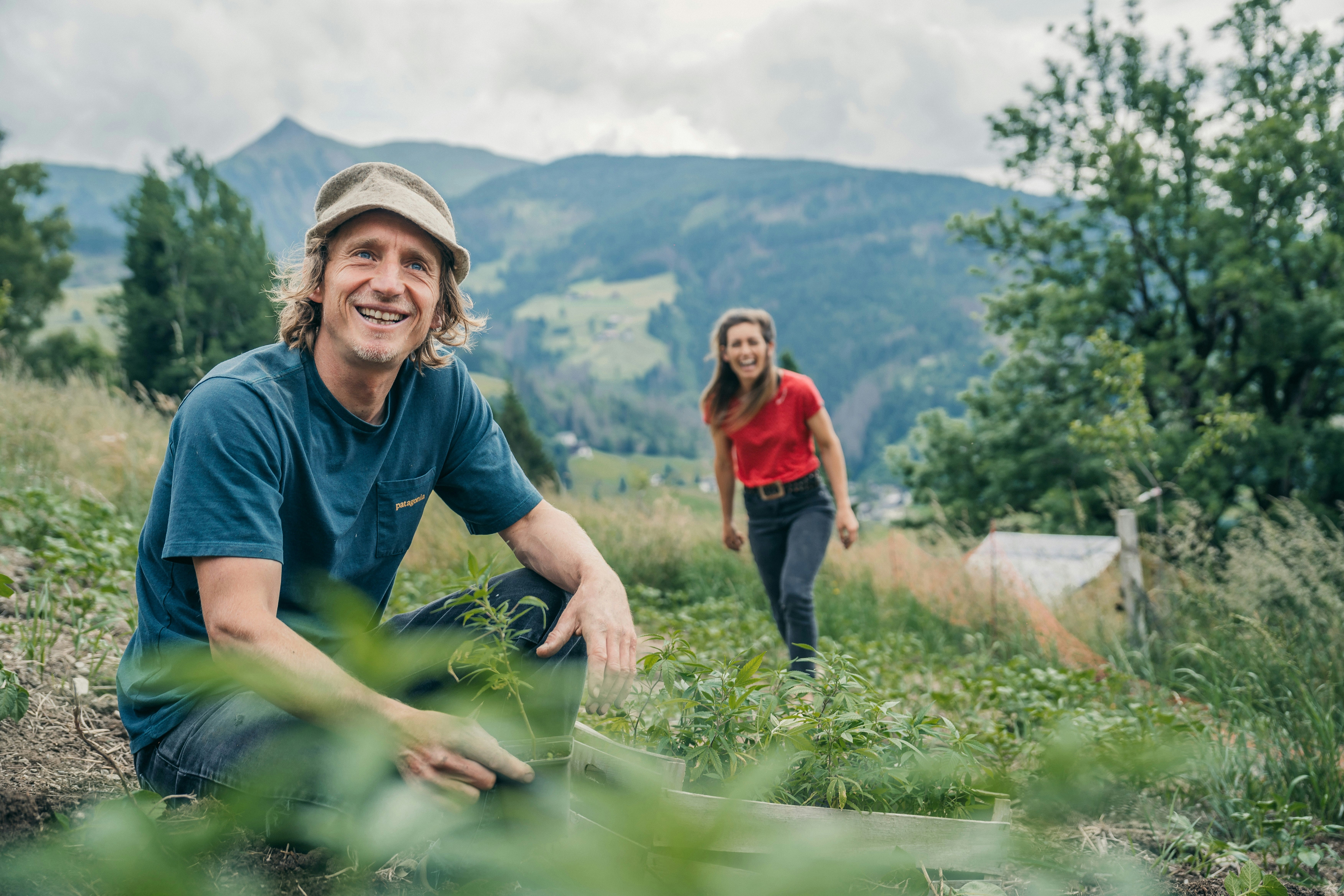 Ein Camper und eine Frau laufen über eine Wiese am Campingplatz, im Hintergrund Berge und Bäume.