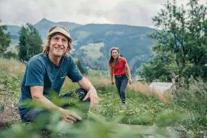 Ein Camper und eine Frau laufen über eine Wiese am Campingplatz, im Hintergrund Berge und Bäume.
