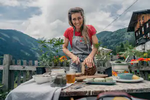 Frühstückstisch auf einer Camping-Terrasse mit Blumen, Getränkeglas und Blick auf Berge und Holzhäuser.