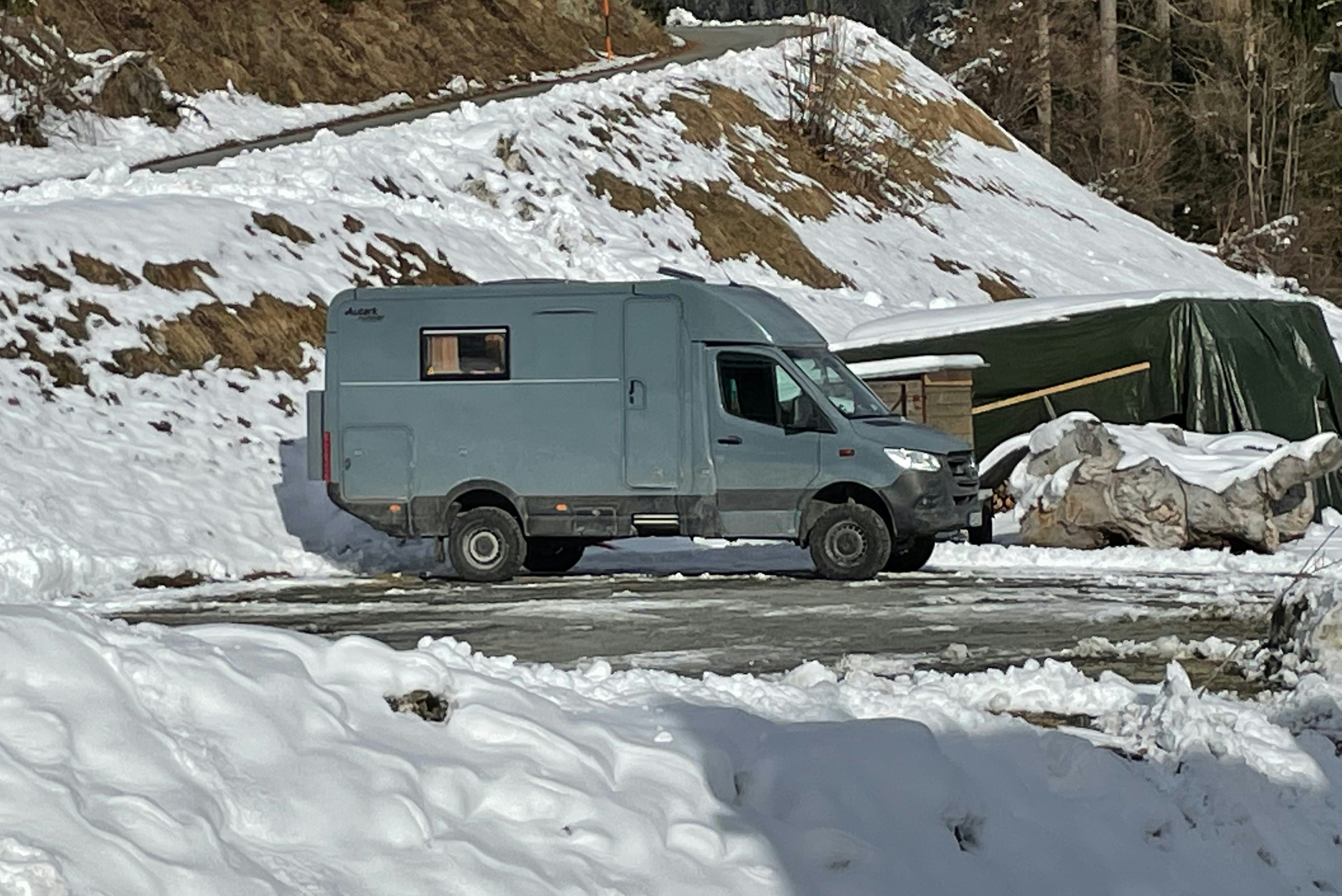 Wohnmobil im Schnee auf einem Campingplatz, im Hintergrund verschneite Berge und Nadelbäume.