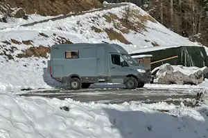 Wohnmobil im Schnee auf einem Campingplatz, im Hintergrund verschneite Berge und Nadelbäume.