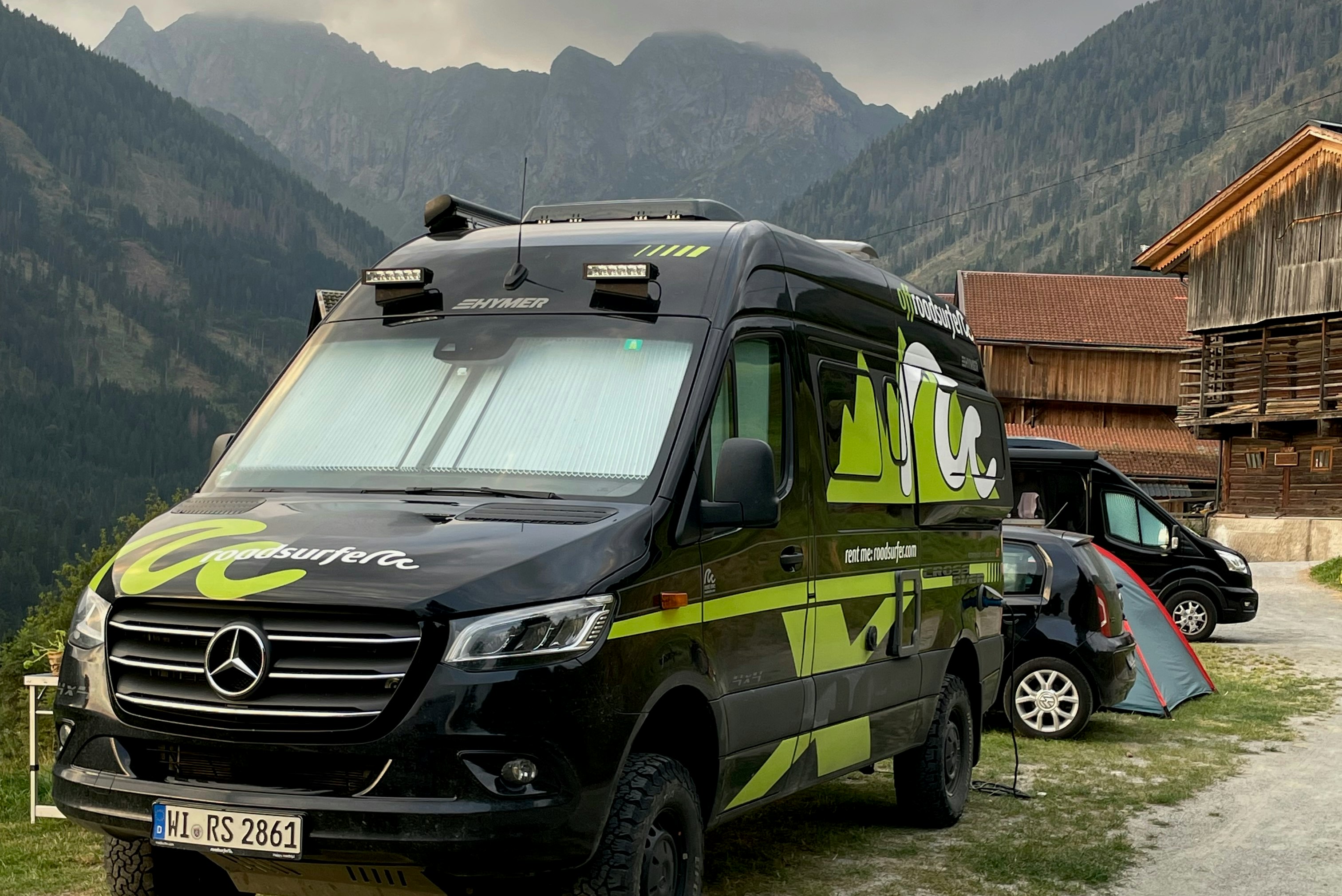 Grünes Wohnmobil mit Markise am Campingplatz, im Hintergrund Berge und ein Holzgebäude.