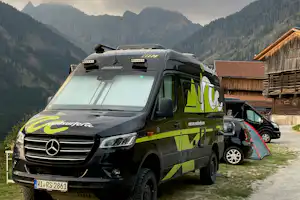 Grünes Wohnmobil mit Markise am Campingplatz, im Hintergrund Berge und ein Holzgebäude.