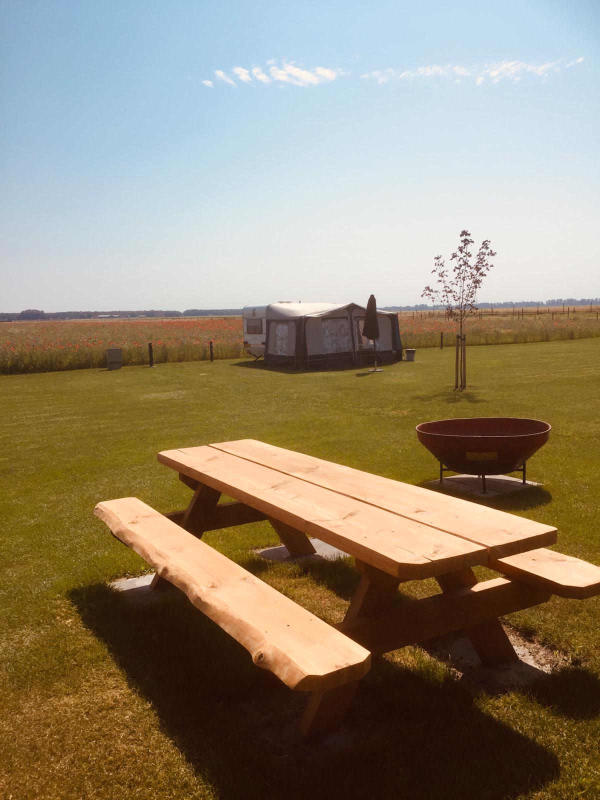 Picknicktisch auf einer grünen Wiese vor einem Zelt und einem Wohnwagen, im Hintergrund Felder und freier Himmel.
